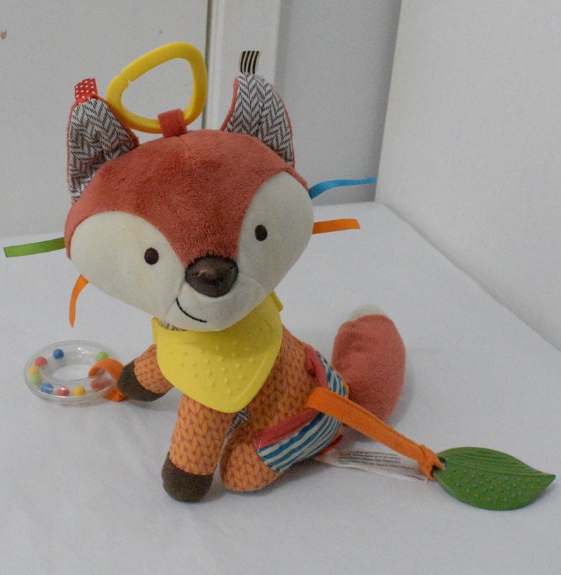 May include: A plush fox baby toy with a yellow bib, orange and white patterned body, and a leaf-shaped teether. The toy has a yellow ring for hanging and colorful ribbons. A small rattle is also included.