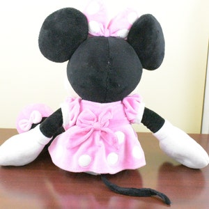 May include: A plush Minnie Mouse doll, viewed from the back, features a black head with large ears and a pink polka-dot bow. She wears a pink dress with white polka dots and a matching bow. The doll has white gloves and a black tail.