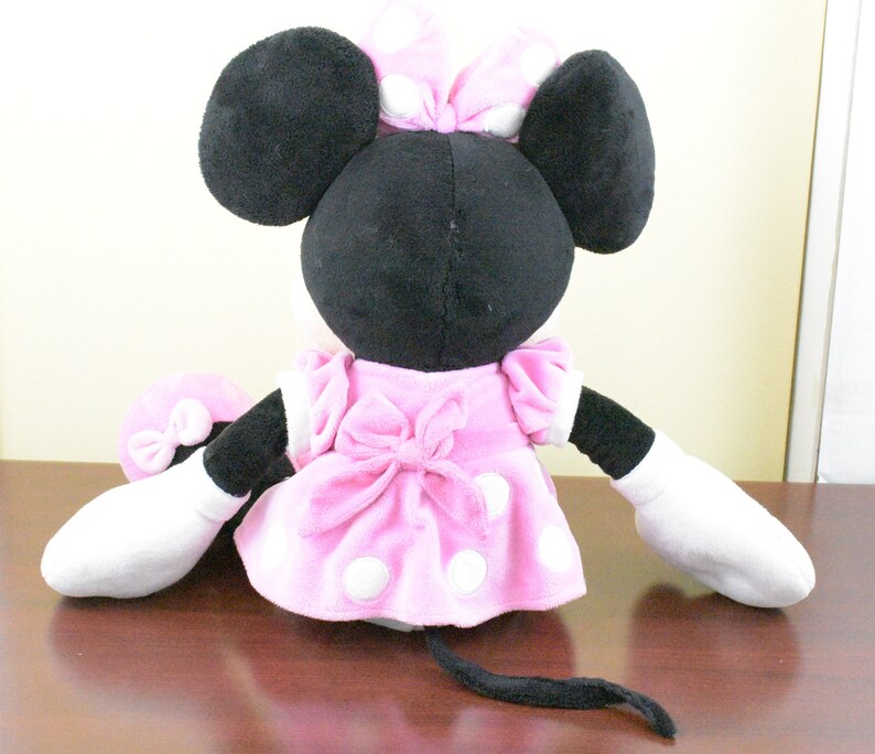 May include: A plush Minnie Mouse doll, viewed from the back, features a black head with large ears and a pink polka-dot bow. She wears a pink dress with white polka dots and a matching bow. The doll has white gloves and a black tail.
