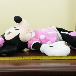 May include: A large Minnie Mouse plush toy, primarily black with white gloves, a pink dress with white polka dots, and pink shoes. The toy is approximately 42 inches long, lying on a brown surface.