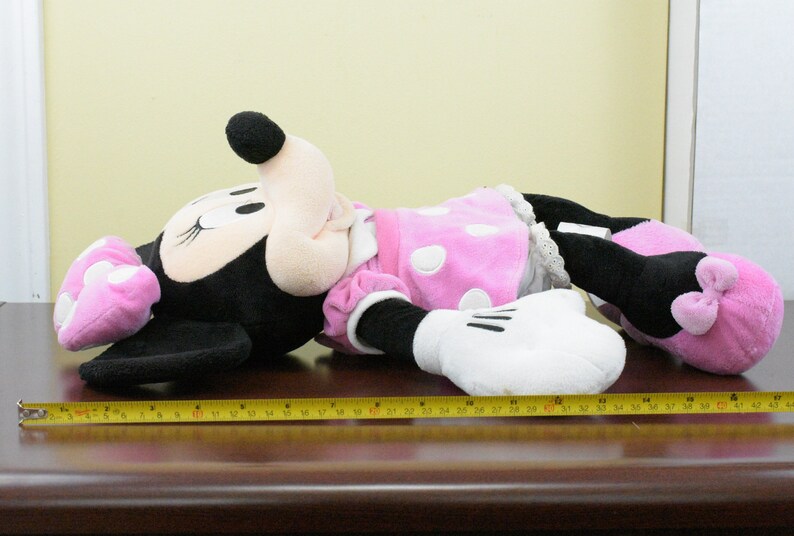 May include: A large Minnie Mouse plush toy, primarily black with white gloves, a pink dress with white polka dots, and pink shoes. The toy is approximately 42 inches long, lying on a brown surface.