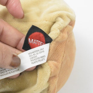 May include: A close-up shot of a plush toy in tan and cream hues. A black tag with the red "MATTEL" logo is visible, alongside a white tag detailing "POLYESTER FIBRES" and "PLASTIC PELLETS" as contents. The soft texture of the toy is clearly shown.