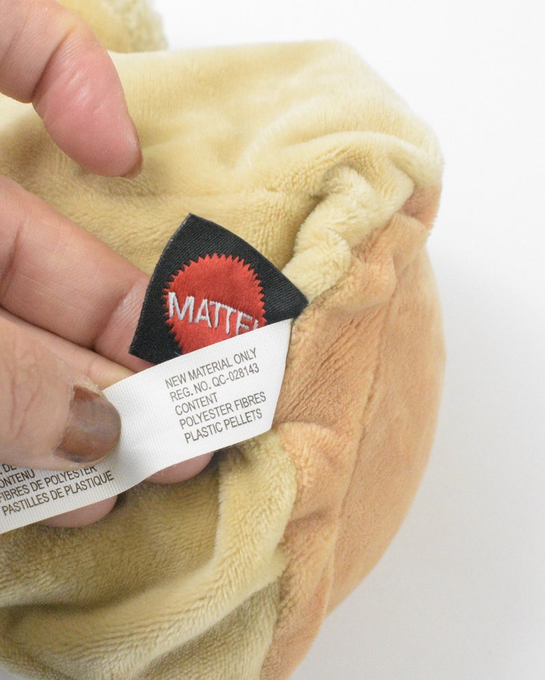 May include: A close-up shot of a plush toy in tan and cream hues. A black tag with the red "MATTEL" logo is visible, alongside a white tag detailing "POLYESTER FIBRES" and "PLASTIC PELLETS" as contents. The soft texture of the toy is clearly shown.