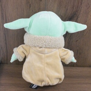 May include: A plush toy of a character from the Star Wars series, with large ears and a tan robe. The toy has a light green head and ears, with a cream-coloured, fuzzy collar. The back of the toy is visible.