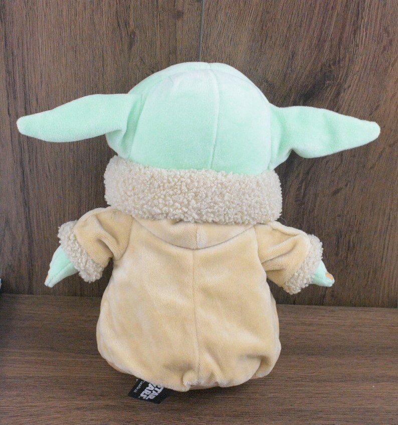 May include: A plush toy of a character from the Star Wars series, with large ears and a tan robe. The toy has a light green head and ears, with a cream-coloured, fuzzy collar. The back of the toy is visible.