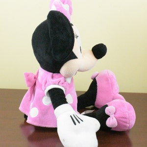 May include: A plush Minnie Mouse doll in a seated position. The doll features a pink dress with white polka dots, a matching bow, and a pink bow on her head. Her black ears and nose are prominent, and she wears white gloves.