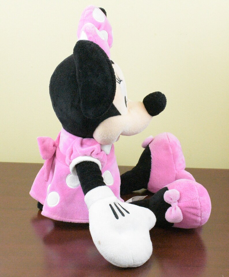 May include: A plush Minnie Mouse doll in a seated position. The doll features a pink dress with white polka dots, a matching bow, and a pink bow on her head. Her black ears and nose are prominent, and she wears white gloves.