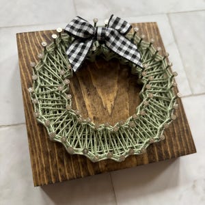 May include: A handmade string art wreath in a circular design, crafted with light green thread and secured with small nails on a stained wooden square. A black and white checkered bow is attached at the top. The wreath is a decorative item.