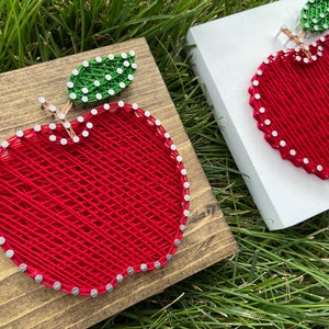May include: Two string art apple designs on wooden plaques. One apple is on a brown plaque and the other is on a white plaque. Both apples are red with green leaves.