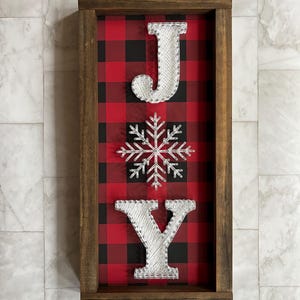 以下が含まれることがあります： 赤と黒のバッファローチェックの背景に、白いストリングアートで「JOY」の文字が書かれた木製フレームのサイン。白い雪の結晶が文字の間に配置されています。
