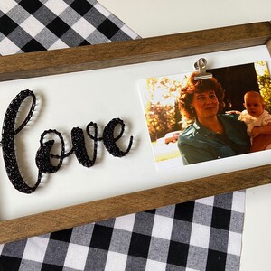 May include: A wooden frame with a white background. The word "love" is spelled out in black string art. A photo of a woman and a baby is clipped to the frame with a silver clip.