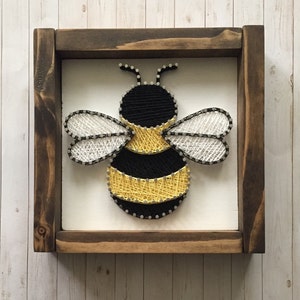 May include: A framed string art piece depicting a bumblebee with black and yellow stripes, white wings, and a black head. The frame is made of dark wood.