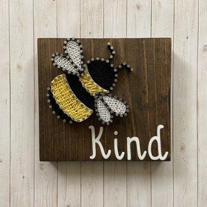 May include: A wooden sign with a string art bee design in yellow, black, and white. The sign says "Kind" in white lettering.