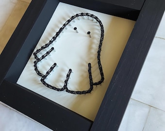 Puede incluir: Una pieza de arte de hilo enmarcada con un diseño de fantasma blanco. El fantasma está delineado con hilo negro y asegurado con pequeñas uñas sobre un fondo blanco. El marco es negro.