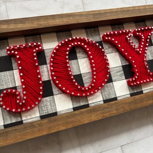 以下が含まれることがあります： 「JOY」の文字が赤い糸で書かれた木製のフレーム付きサイン。文字は小さな銀色のスタッドで縁取られ、白黒のチェック柄の背景に配置されています。