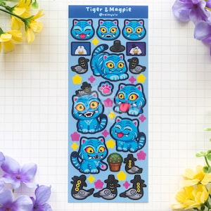 Può includere: Un foglio di adesivi con illustrazioni di gatti e uccelli blu a cartoni animati. I gatti hanno varie espressioni e indossano cappelli. Il testo "Tiger & Magpie" è in alto. Lo sfondo è bianco con un motivo a griglia, e ci sono fiori.
