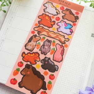 May include: A sticker sheet with cartoon horse designs in various colours and poses. The stickers include a white horse, a brown horse, an orange horse with a fiery mane, and a pink pegasus. The sheet is on a pink background with the word "HORSES!" at the top.