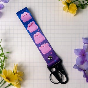 May include: A blue and purple lanyard featuring pink cartoon characters. The lanyard has a black metal clasp and key ring. The background is a grid pattern with flowers.