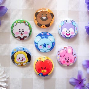 May include: A collection of seven colorful, round enamel pins featuring cartoon characters. The pins are in various colors, including green, orange, blue, purple, yellow, red, and pink. The background is a checkered pattern with purple flowers.