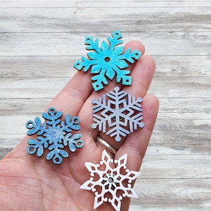 May include: Four wooden snowflake decorations in shades of blue and white are shown. The snowflakes have detailed cut-out patterns and glitter highlights. The decorations are held in a hand against a light wood background.