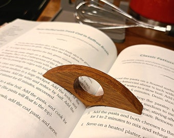 May include: A wooden page holder rests on an open cookbook. The page holder is made of light brown wood with a circular opening. The cookbook is open to a recipe with text. A whisk and red pot are in the background.