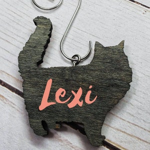 May include: A wooden cat-shaped ornament with the name "Lexi" in coral lettering. The ornament is dark gray with a silver metal hanger. The cat silhouette has a fluffy tail and is set against a light wood grain background.