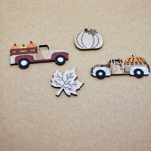 May include: Wooden fall-themed cutouts including two trucks carrying pumpkins, a pumpkin, and a maple leaf. The trucks are brown, red, and white. The pumpkins are orange and yellow. The leaf and pumpkin have green accents.