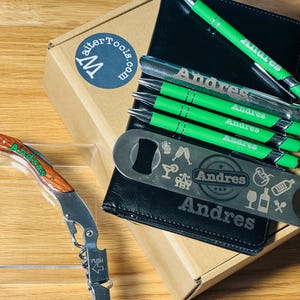 May include: A black leatherette gift box with a personalized bottle opener and four green pens with the name "Andres" engraved on them. The box is open and a wooden corkscrew is visible in the foreground.