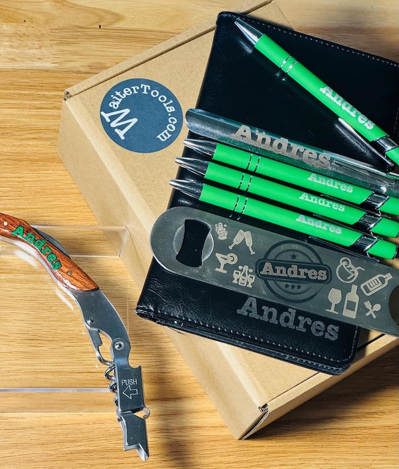 Puede incluir: Una caja de regalo de imitaci&oacute;n de cuero negro con un abridor de botellas personalizado y cuatro bol&iacute;grafos verdes con el nombre "Andres" grabado en ellos. La caja est&aacute; abierta y se puede ver un sacacorchos de madera en primer plano.