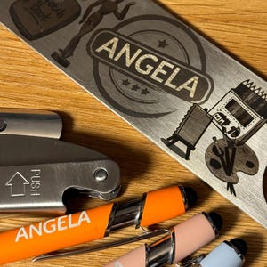 Puede incluir: Tres bol&iacute;grafos personalizados con el nombre "Angela" grabado en ellos. Los bol&iacute;grafos son naranja, rosa y azul. Los bol&iacute;grafos est&aacute;n al lado de un abridor de botellas de metal con el nombre "Angela" grabado en &eacute;l. El abridor de botellas tiene un dise&ntilde;o de un cuaderno de bocetos, un par de tijeras, un maniqu&iacute; y una paleta de pintura.
