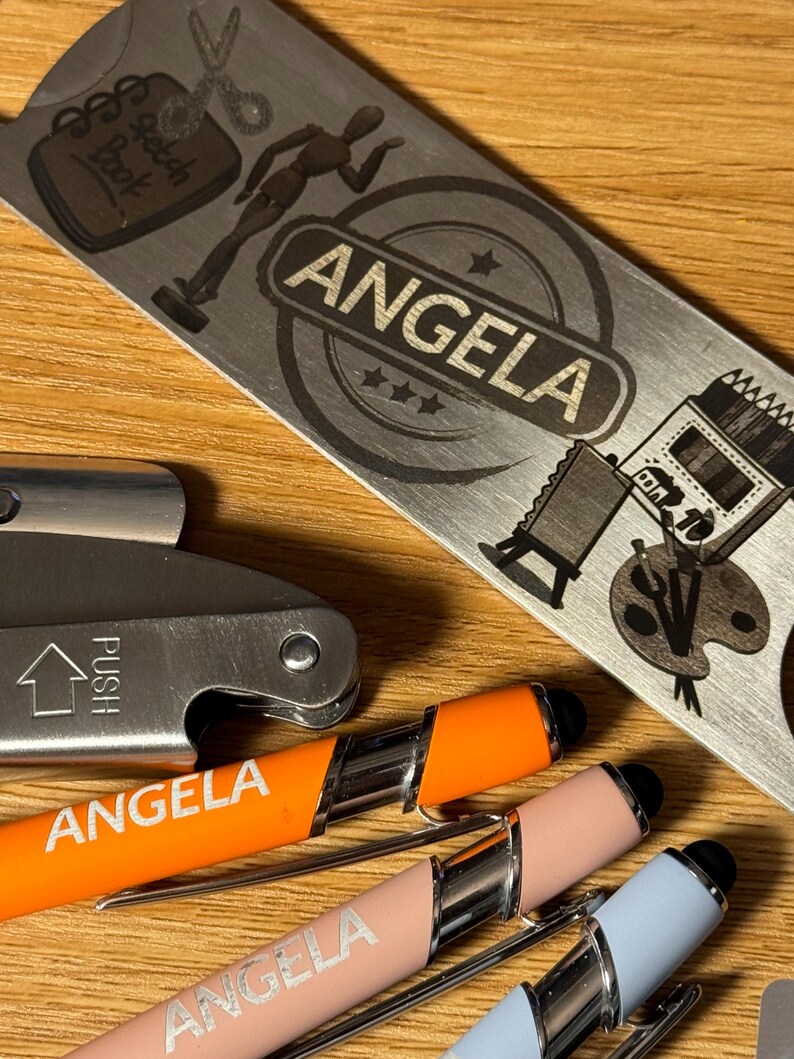 Puede incluir: Tres bol&iacute;grafos personalizados con el nombre "Angela" grabado en ellos. Los bol&iacute;grafos son naranja, rosa y azul. Los bol&iacute;grafos est&aacute;n al lado de un abridor de botellas de metal con el nombre "Angela" grabado en &eacute;l. El abridor de botellas tiene un dise&ntilde;o de un cuaderno de bocetos, un par de tijeras, un maniqu&iacute; y una paleta de pintura.