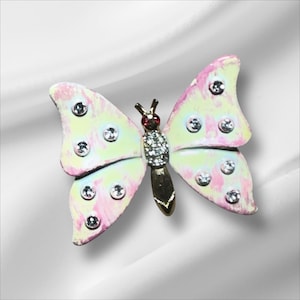 May include: A butterfly brooch with pink, yellow, and white wings. The brooch has a gold-colored body and is adorned with clear, round rhinestones and a red gem. The wings have a marbled effect.