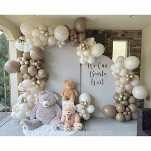 May include: A white backdrop with the text "We Can Bearly Wait" decorated with a balloon arch made of brown, beige, and gold balloons. Three teddy bears are in front of the backdrop.