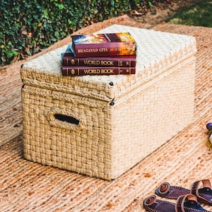 May include: A woven wicker storage chest with a lid, sitting on a woven rug. Three books are stacked on top of the chest, including "Bhagavad Gita As It Is", "World Book", and "World Book".