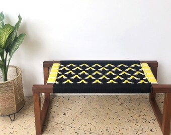 May include: A black and yellow woven bench with a wooden frame. The bench has a geometric pattern and is sitting on a light brown tiled floor.