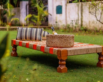 May include: A wooden outdoor daybed with a woven jute mattress and red and yellow stripes. A black and white striped pillow is on the daybed. A woven basket with a bottle of wine is on the daybed.