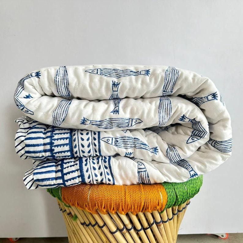 Puede incluir: Edred&oacute;n plegado blanco y azul estampado con dise&ntilde;os de peces y geom&eacute;tricos. El edred&oacute;n est&aacute; apilado sobre un taburete tejido con tonos naranja, verde y naturales. El dise&ntilde;o del edred&oacute;n incluye peces y otras formas.