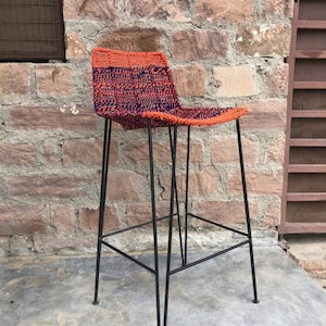 May include: A black metal bar stool with a woven seat in orange and blue. The stool has a simple design with a single bar across the back.