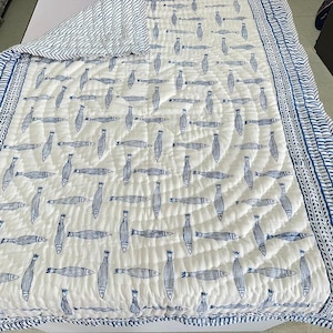 Puede incluir: Colcha blanca y azul estampada con dise&ntilde;o de peces. La colcha presenta un patr&oacute;n repetitivo de peces azules sobre un fondo blanco. El borde es un dise&ntilde;o de rayas azules y blancas. La colcha est&aacute; hecha de un material acolchado suave.