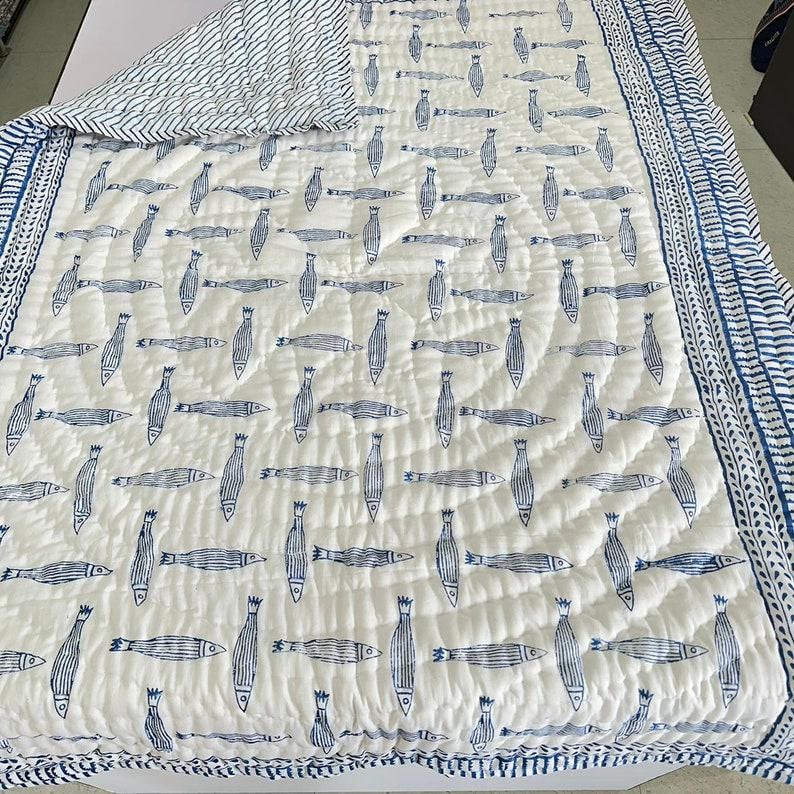 Puede incluir: Colcha blanca y azul estampada con dise&ntilde;o de peces. La colcha presenta un patr&oacute;n repetitivo de peces azules sobre un fondo blanco. El borde es un dise&ntilde;o de rayas azules y blancas. La colcha est&aacute; hecha de un material acolchado suave.