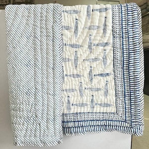 Puede incluir: Un edredón blanco y azul con un estampado de peces. El edredón presenta un panel central con motivos de peces azules sobre un fondo blanco, bordeado por un diseño de rayas azules y blancas. El lado izquierdo tiene un patrón de rayas diagonales.