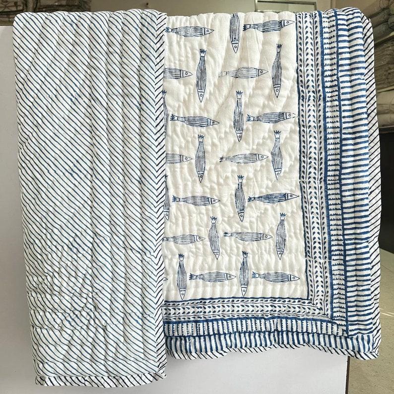 Puede incluir: Un edred&oacute;n blanco y azul con un estampado de peces. El edred&oacute;n presenta un panel central con motivos de peces azules sobre un fondo blanco, bordeado por un dise&ntilde;o de rayas azules y blancas. El lado izquierdo tiene un patr&oacute;n de rayas diagonales.