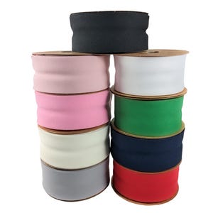 May include: A stack of nine spools of fabric ribbon in various solid colors including black, light pink, bright pink, cream, gray, white, green, navy blue, and red. Each spool features a central horizontal indentation along the ribbon surface.