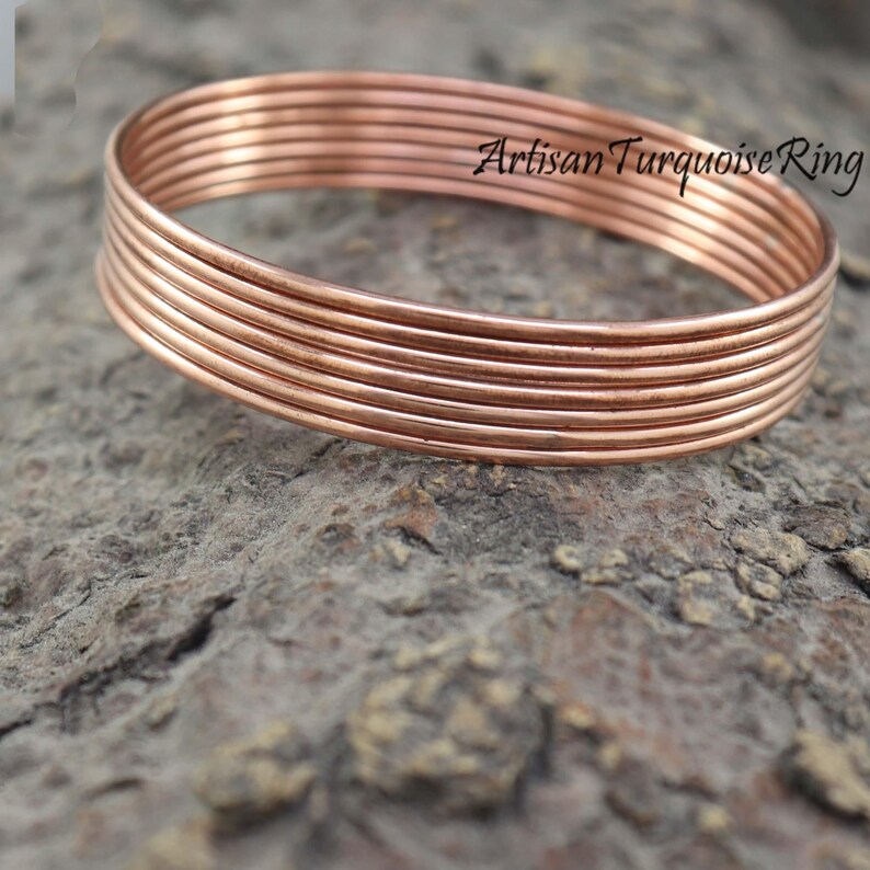 May include: A stack of copper-colored bangles, with the text "Artisan Turquoise Ring" in black script. The bangles are stacked on a textured, gray-brown surface. The jewelry is made of thin, round metal.