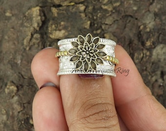 Puede incluir: Un anillo plateado con un diseño de flor dorada. El anillo tiene una banda texturizada y una banda dorada alrededor de la flor. El anillo está en un dedo.