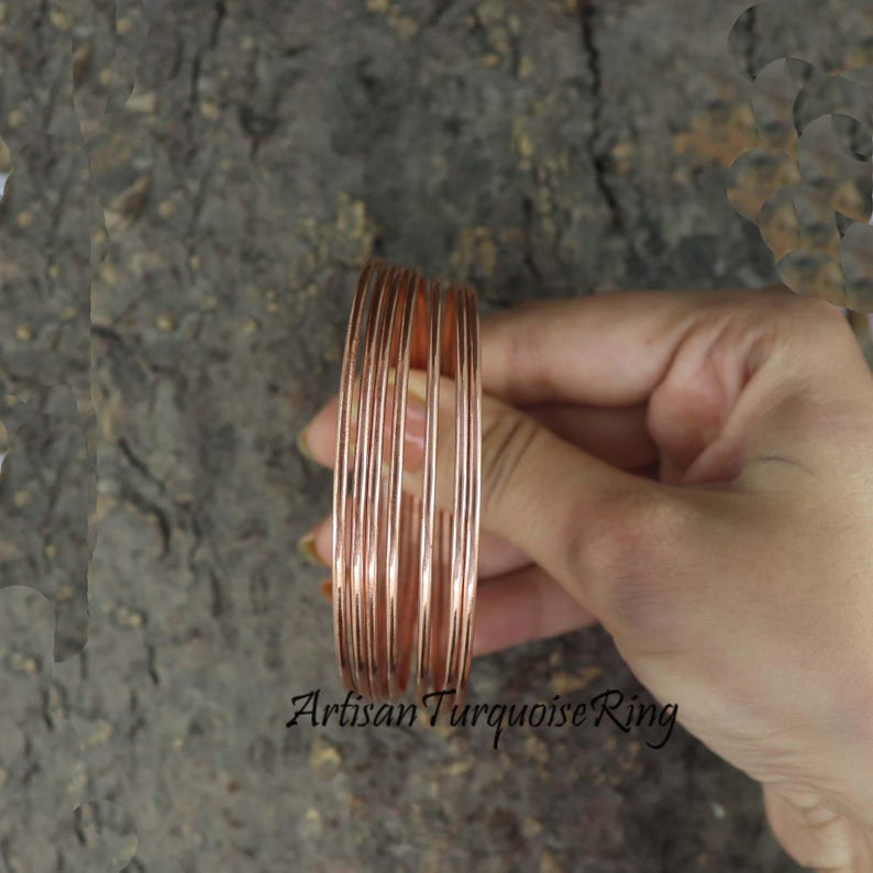 May include: A stack of copper-colored bangles. The thin, round bracelets are held in a hand, with the Artisan Turquoise Ring logo visible at the bottom. The bangles are a warm, metallic color.