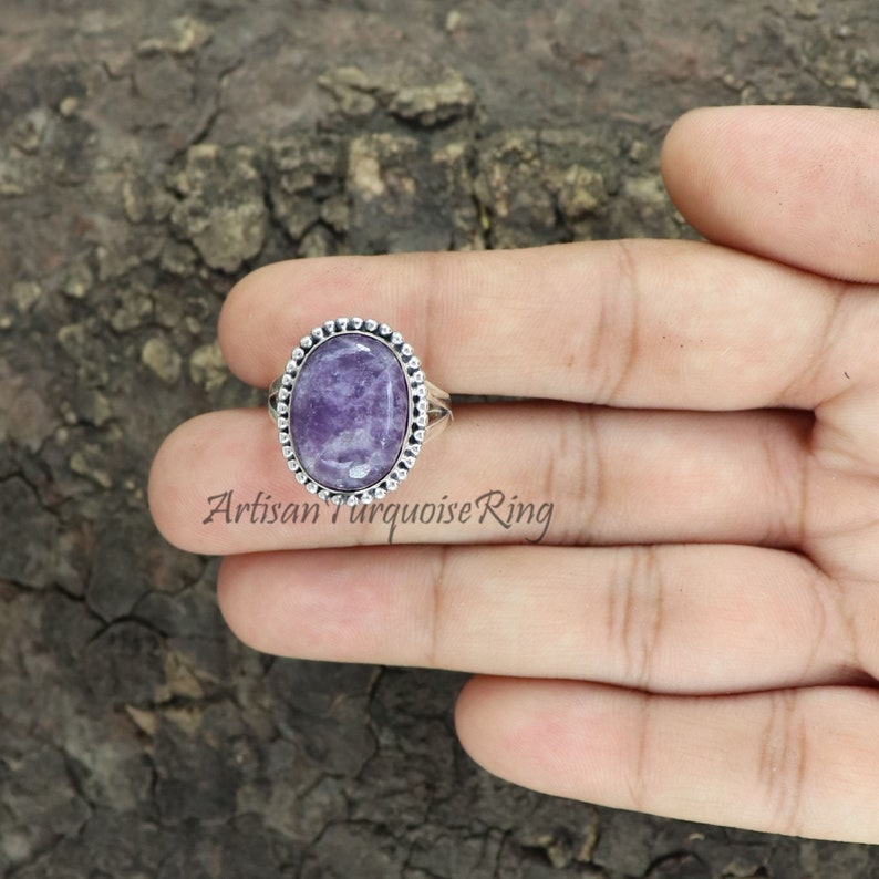 K&ouml;nnte beinhalten: Ein silberner Ring mit einem ovalen, violetten Edelstein, der in einer perlenbesetzten Fassung sitzt. Der Ring befindet sich an einem Finger.