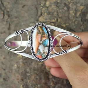 May include: A silver cuff bracelet with a large oval-shaped turquoise stone set in the center. The stone has a mix of blue, green, orange, and white colors. The bracelet has a delicate, intricate design.