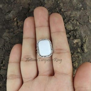 May include: A white gemstone ring with a beaded silver setting. The ring is on a person's finger.