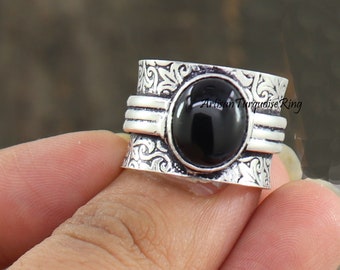 Puede incluir: Un anillo plateado con una piedra negra ovalada grande en el centro. El anillo tiene un diseño grabado y adornado en la banda y el texto "Artisan Turquoise Ring". El anillo está sostenido por una persona.